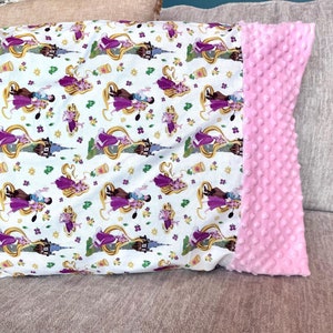 May include: A white pillow with a pink minky back. The front of the pillow features a repeating pattern of Rapunzel and Flynn Rider from the Disney movie Tangled.