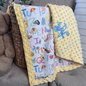 May include: A yellow and white baby blanket with a colorful alphabet print and a blue embroidered name "Nathan" on the yellow side.