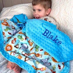 May include: A blue and white baby blanket with a  pattern of animals, including giraffes, elephants, monkeys, and lions. The blanket has a blue minky backing and is personalized with the name "Blake".