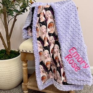 May include: A lavender baby blanket with a soft, textured surface and a printed interior featuring cartoon characters. The blanket has the name "Emory Rose" embroidered in pink on the corner. The blanket is folded over a small wooden stool.