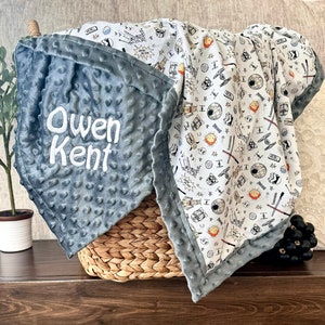 May include: A gray and white baby blanket with a Star Wars theme. The blanket has a gray minky side and a white cotton side with a print of Star Wars characters and symbols. The blanket is personalized with the name "Owen Kent" embroidered on the gray side.