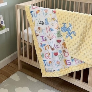 May include: A baby blanket featuring a white fabric side with a colorful alphabet print including various animals and objects, and a solid pale yellow dimpled minky fabric side. The yellow side displays the embroidered name "Nala" in blue script.