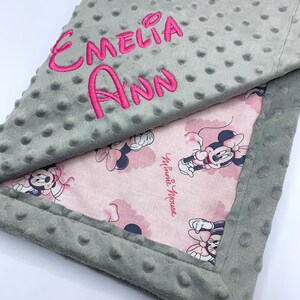 May include: A gray and pink baby blanket with a Minnie Mouse print and the name "Emeli Ann" embroidered on the gray side.