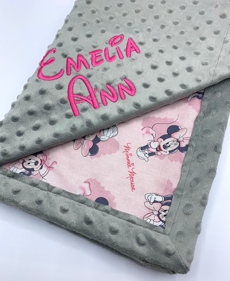 May include: A gray and pink baby blanket with a Minnie Mouse print and the name "Emeli Ann" embroidered on the gray side.