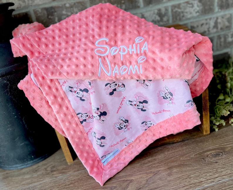 May include: A pink and white baby blanket with a Minnie Mouse print on one side and the name "Sophia Naomi" embroidered on the other side.