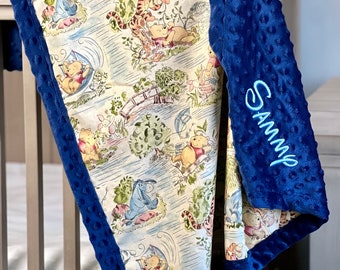 May include: A Winnie the Pooh themed baby blanket with a navy blue minky backing. The blanket features a light cream background with a repeating pattern of Winnie the Pooh, Piglet, Tigger, and Eeyore characters. The blanket is personalized with the name "Sammy" embroidered in blue on the navy blue backing.
