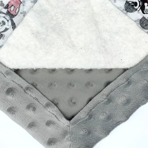 May include: Close-up of a gray and white baby blanket with a Mickey Mouse design. The blanket is made of soft, plush fabric and has a white layer of batting in the middle.