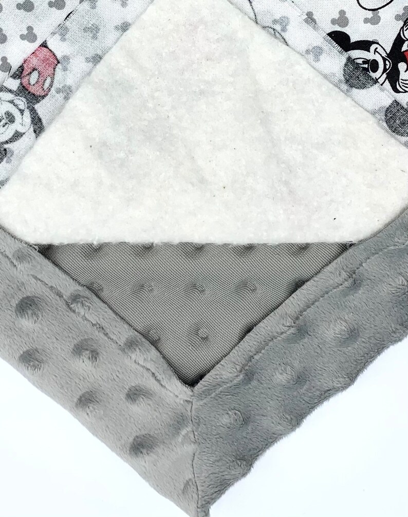 May include: Close-up of a gray and white baby blanket with a Mickey Mouse design. The blanket is made of soft, plush fabric and has a white layer of batting in the middle.