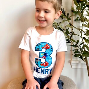 Op de afbeelding: Een wit T-shirt met een blauw nummer drie op de voorkant. Het nummer drie is versierd met kleurrijke vissen en bubbels. De naam "HENRY" staat in blauwe letters onder het nummer. Een jong kind draagt het shirt en glimlacht.