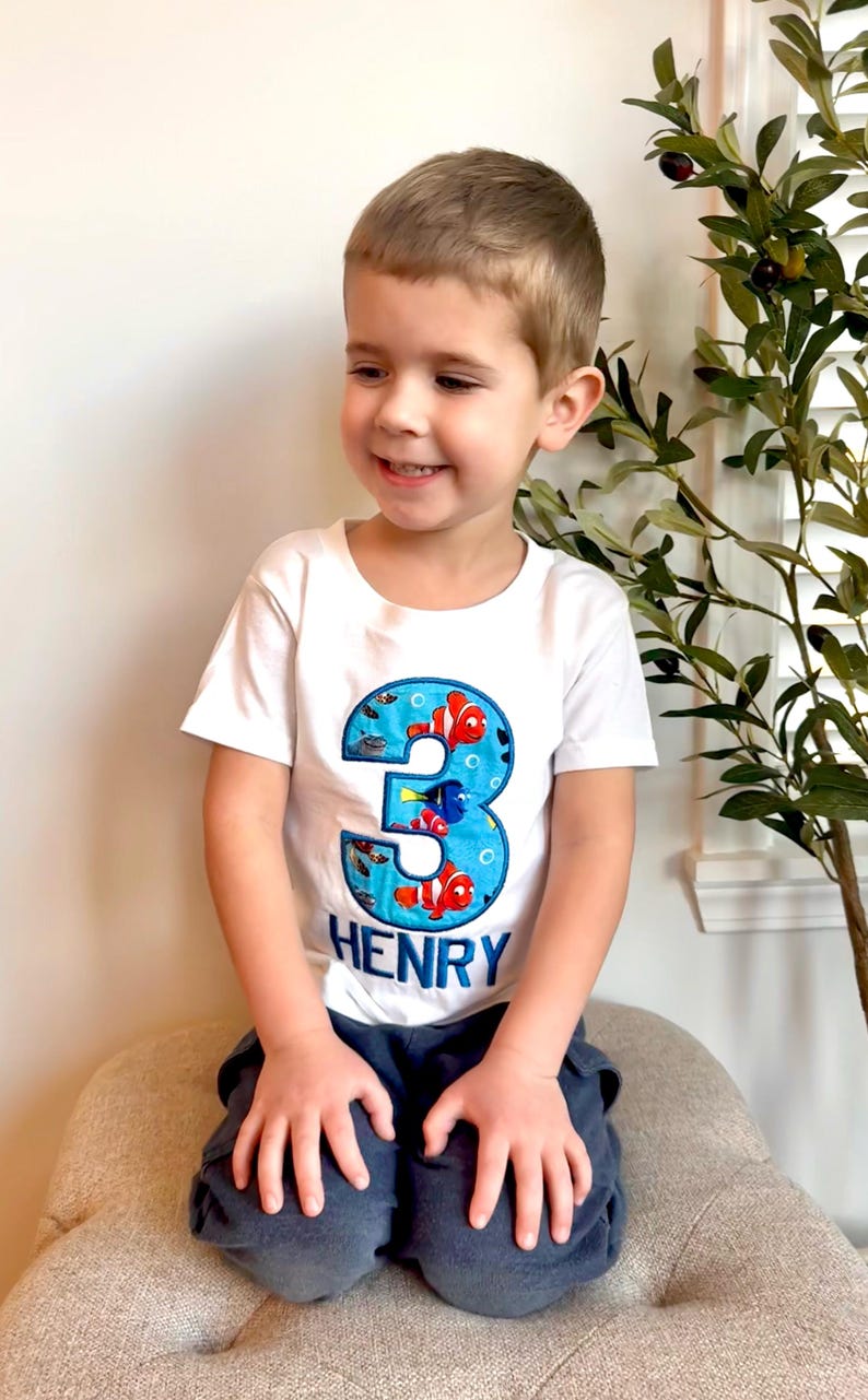 Op de afbeelding: Een wit T-shirt met een blauw nummer drie op de voorkant. Het nummer drie is versierd met kleurrijke vissen en bubbels. De naam "HENRY" staat in blauwe letters onder het nummer. Een jong kind draagt het shirt en glimlacht.