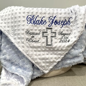May include: White and light blue baby blanket with embroidered text "Blake Joseph Baptized in Christ August 25th 2024" and a cross.