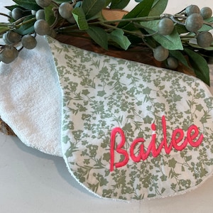 May include: A white and green floral print burp cloth with a pink embroidered name "Bailee" on the front. The cloth is made of soft, absorbent material and has a white terry cloth backing.