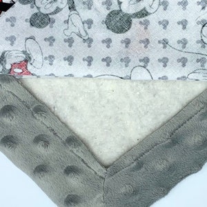 May include: A gray and white baby blanket with a Mickey Mouse print. The blanket has a soft, plush texture and is perfect for cuddling.