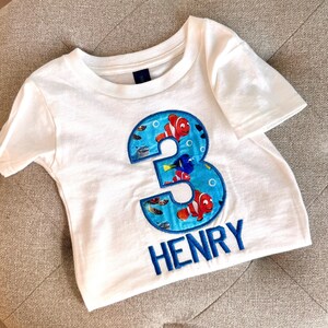 Op de afbeelding: Wit T-shirt met een blauw nummer 3, met een Finding Nemo-thema met kleurrijke vissen en bubbels. De naam "HENRY" is geborduurd onder het nummer.