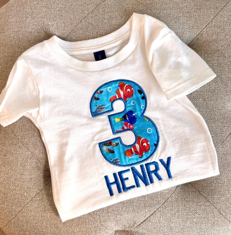 Op de afbeelding: Wit T-shirt met een blauw nummer 3, met een Finding Nemo-thema met kleurrijke vissen en bubbels. De naam "HENRY" is geborduurd onder het nummer.