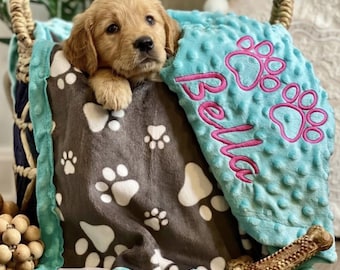 May include: A golden retriever puppy resting inside a woven basket draped with a soft blanket. The blanket features a dark gray section with white paw prints and a textured light blue section embroidered with the name Bella and pink paw prints.