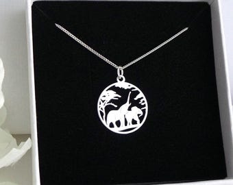 Puede incluir: Un collar plateado con un colgante circular que presenta una silueta de elefante y jirafa en un paisaje. El colgante cuelga de una fina cadena, presentado en una caja blanca con interior negro.