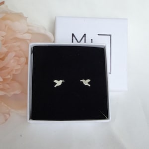 May include: Silver bird stud earrings presented in a black box with a white lid. The earrings are small and delicate, featuring a minimalist design. The box is open, showcasing the earrings against a black backdrop.