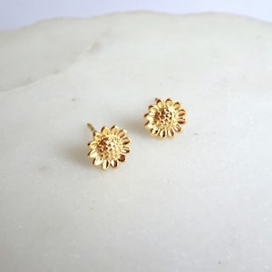 May include: A pair of gold-toned stud earrings shaped like sunflowers. Each earring features a textured, raised centre surrounded by a ring of small, rounded petals. The earrings have a polished metallic finish and include a standard post backing.