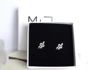 May include: A pair of silver-toned snake-shaped stud earrings displayed inside a square white box with a black velvet insert. The box lid is white with the black text M.J visible on the front. The earrings feature a textured, winding snake design.