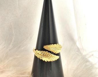 May include: A gold-colored ring with a feather design. The ring wraps around the finger with two detailed feather motifs. The ring is displayed on a black cone-shaped stand against a backdrop of white feathers.