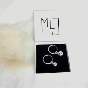 May include: Two silver rings with a dangling silver ball charm. The rings are in a black box with a white lid. The lid has the text "ML" and "Me Lou Jewellery" printed on it.