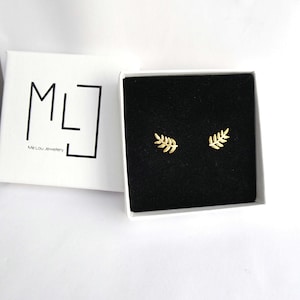 May include: A pair of gold-coloured leaf stud earrings presented in an open white box with a black interior. The box is branded with the text "ML" and "Me Lou Jewelry".