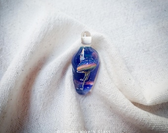 May include: A handcrafted glass pendant with a jellyfish design. The pendant is a deep blue color with a jellyfish in the center, featuring pink, yellow, and white hues. The pendant has a clear stopper and is suspended on a white cord.