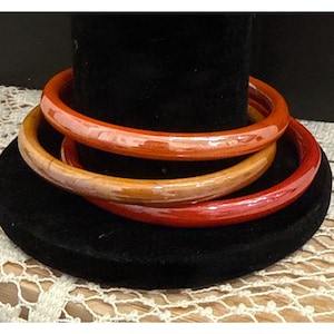 May include: Three wooden bangles in shades of red, orange, and tan are displayed on a black velvet stand. The bangles are smooth and glossy, showcasing the natural wood grain. The stand sits on a white lace doily.