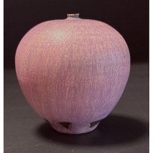 May include: A round, lavender-colored ceramic vase with a textured surface. The vase has a small opening at the top and three triangular cutouts at the base. The color is a blend of purple and beige.