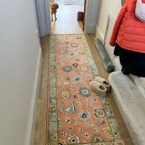 May include: A long, rectangular rug with a peach background and a floral pattern in shades of blue, green, and cream. The rug is placed in a hallway with wooden flooring. A pair of fuzzy slippers and a dog are also visible.