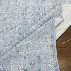 Puede incluir: Alfombra estampada en azul claro y blanco con un aspecto envejecido y vintage. La alfombra presenta un diseño floral detallado y un borde con flecos. La alfombra está parcialmente doblada, revelando la parte inferior.