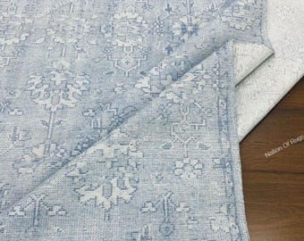 May include: A light blue and white patterned rug with a distressed, vintage look. The rug features a detailed floral design and a fringed edge. The rug is partially folded, revealing the underside.
