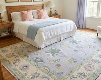 May include: A large rectangular area rug featuring a light blue central field with stylized floral and botanical motifs in muted green, pink, and cream tones. The rug has a wide decorative border with a geometric pattern and white fringe ends.