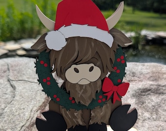 May include: A wooden Highland cow Christmas ornament. The brown cow wears a red Santa hat and a green wreath with red berries. A red bow is on the wreath. The ornament sits on a wooden base and is on a rock.