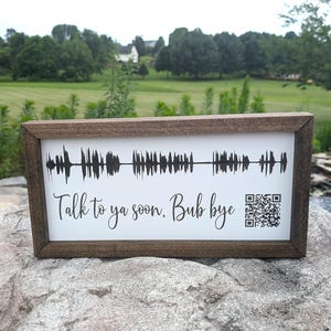 May include: A framed sign with a white background and a brown wooden frame. The sign features a black sound wave design and the text "Talk to ya soon. Bub bye" with a QR code in the lower right corner. The sign is displayed outdoors.