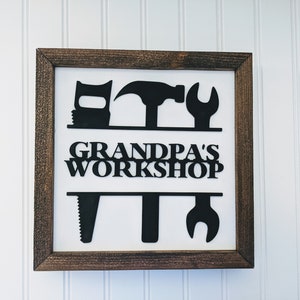 May include: Wooden framed sign with the text "GRANDPA'S WORKSHOP" and black silhouettes of tools. The tools include a saw, wrench, hammer, and other workshop tools. The frame is a dark brown color.