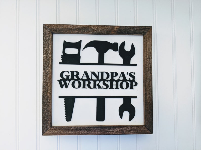 May include: Wooden framed sign with the text "GRANDPA'S WORKSHOP" and black silhouettes of tools. The tools include a saw, wrench, hammer, and other workshop tools. The frame is a dark brown color.