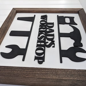 May include: A framed sign with a white background and a dark brown wooden frame. The sign features black cut-out shapes of tools and the words "DAD'S WORKSHOP" in bold, black letters.