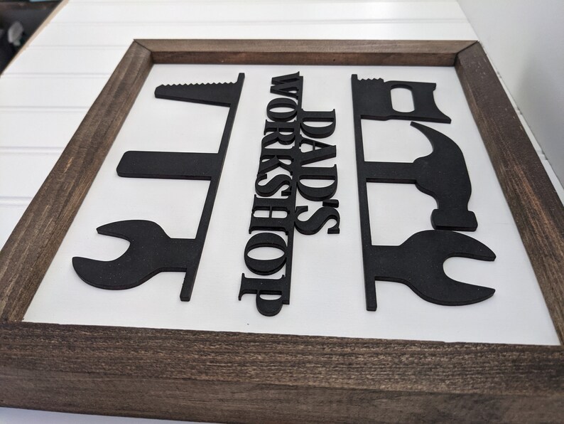 May include: A framed sign with a white background and a dark brown wooden frame. The sign features black cut-out shapes of tools and the words "DAD'S WORKSHOP" in bold, black letters.