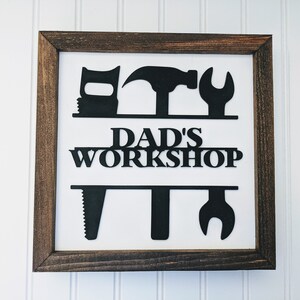 May include: A framed sign with a white background and a dark brown wooden frame. The sign features black silhouettes of tools above and below the words "DAD'S WORKSHOP". The tools include a saw, hammer, and wrench.