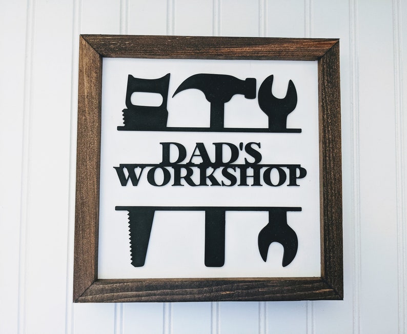 May include: A framed sign with a white background and a dark brown wooden frame. The sign features black silhouettes of tools above and below the words "DAD'S WORKSHOP". The tools include a saw, hammer, and wrench.