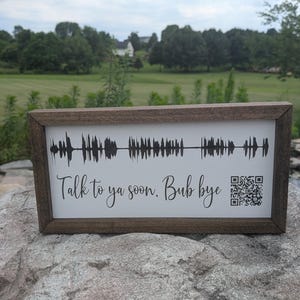 May include: A framed sign with a white background and a black soundwave design. The text "Talk to ya soon. Bub bye" is written below the soundwave. A QR code is in the lower right corner. The frame is brown.