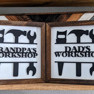 May include: Two framed workshop signs with a white background and brown wooden frames. The signs feature black cut-out tool silhouettes and the text "Grandpa's Workshop" and "Dad's Workshop". Decorative wooden panels are in the background.