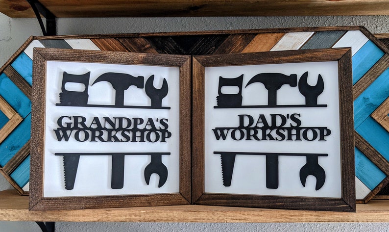 May include: Two framed workshop signs with a white background and brown wooden frames. The signs feature black cut-out tool silhouettes and the text "Grandpa's Workshop" and "Dad's Workshop". Decorative wooden panels are in the background.