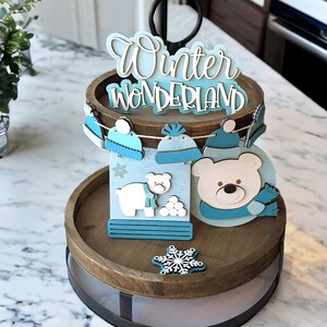 May include: A two-tiered wooden tray with a black handle, decorated with winter-themed cutouts. The top tier features a sign that reads "Winter Wonderland." The decorations include a polar bear, snowballs, and snowflakes. The color scheme is blue, white, and brown.