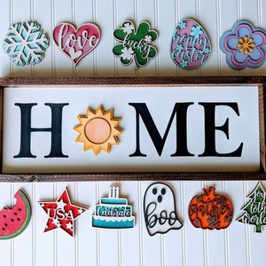 May include: A wooden sign with the word "HOME" in large black letters, with interchangeable seasonal shapes above and below. Shapes include a snowflake, heart, shamrock, Easter egg, flower, watermelon, star, cake, ghost, pumpkin, and Christmas tree.