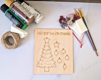 May include: A wooden Christmas tree ornament kit with a laser-cut tree and ornament shapes. Includes paint, brushes, twine, and bows. The text on the wood reads "build your own tree ornament!"