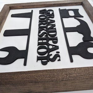 May include: Wooden framed sign with the words "GRANDPA'S WORKSHOP" in black, along with black silhouette cutouts of tools like a saw, wrench, and hammer. The background is white.
