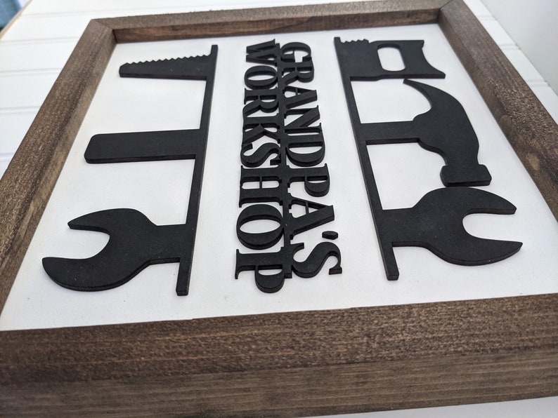 May include: Wooden framed sign with the words "GRANDPA'S WORKSHOP" in black, along with black silhouette cutouts of tools like a saw, wrench, and hammer. The background is white.